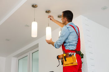 A man in red overalls is installing a light fixture on the ceiling.