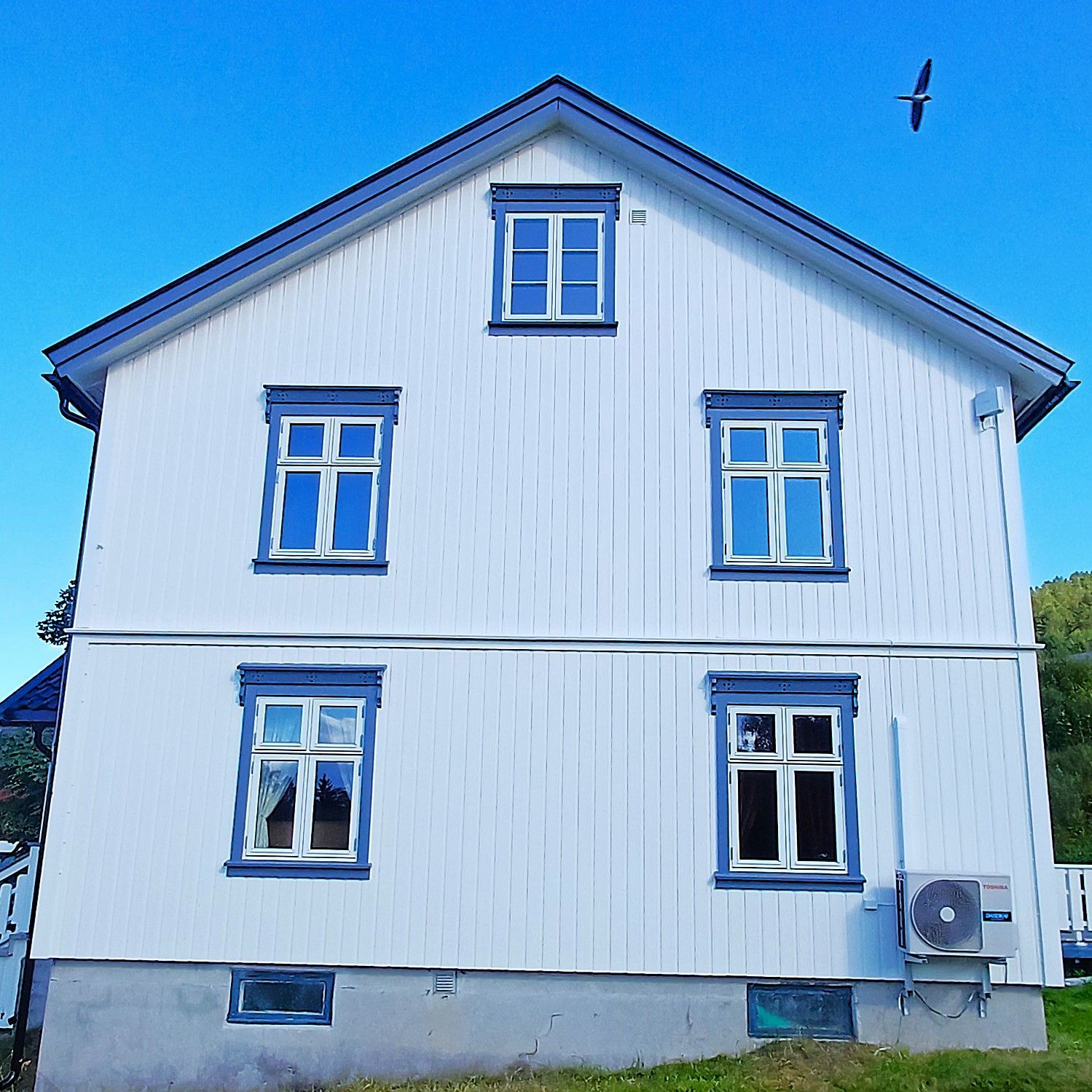 White, two-story house with blue trim and windows against a clear blue sky. A bird flies above.