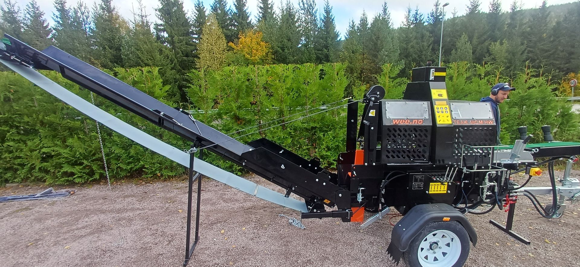A wood splitter with a conveyor belt, on a trailer, in a yard, near greenery.