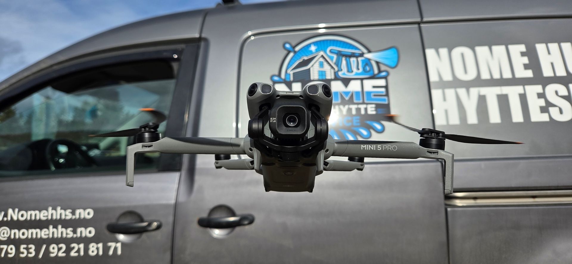 En grå drone svever foran en grå varebil med «Nome Hytteservice»-merking og kontaktinformasjon på siden.