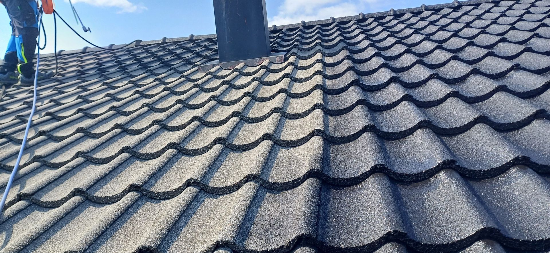 A close-up view of a dark gray tiled roof with a chimney and a person with safety gear.