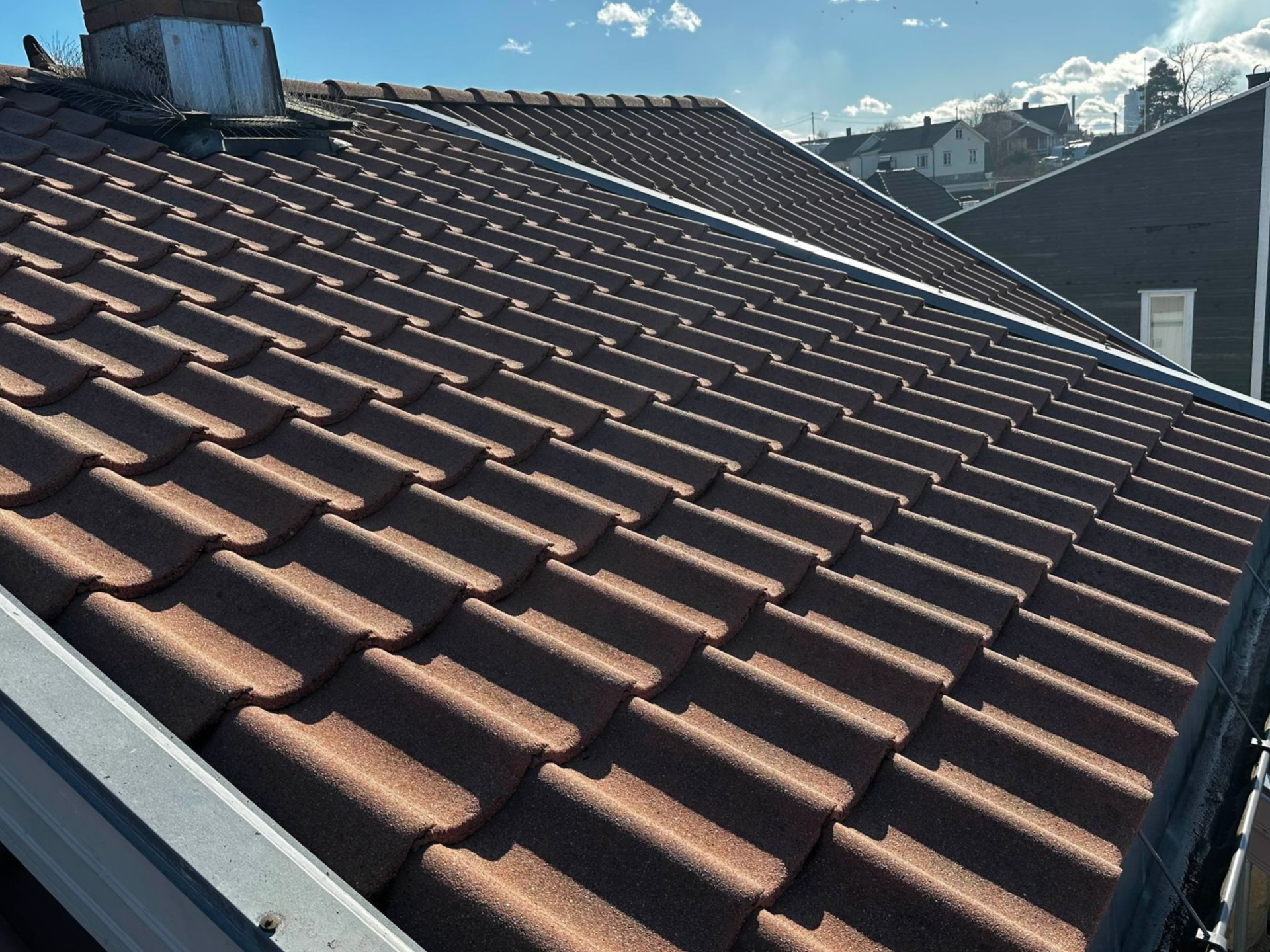 An angled view of a textured, reddish-brown tiled residential roof with a chimney, gutter, and sunny sky in the background.