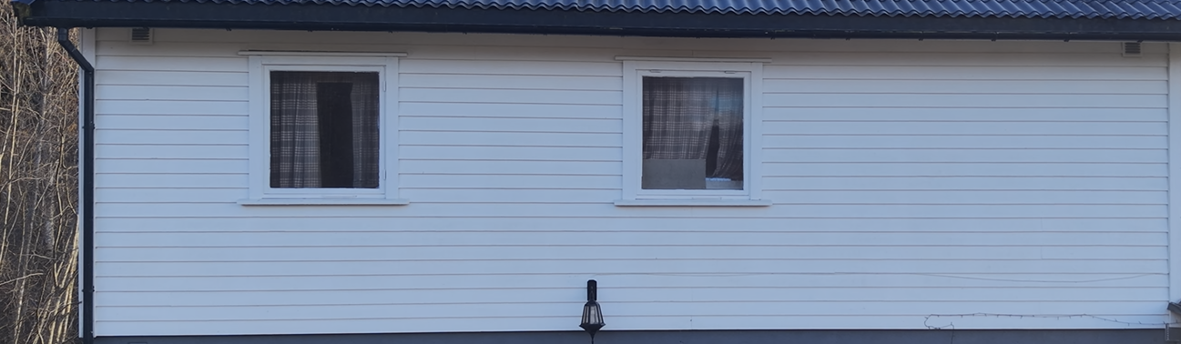 En horisontal seksjon av et hvitt hus med clapboard-paneler, to firkantede vinduer og et svart tak.