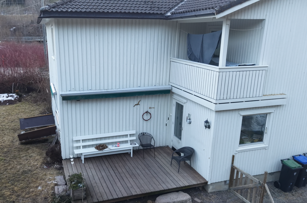 Eksteriøret til et hvitt toetasjes hus med treterrasse, benk, utestoler og en balkong med gardiner i andre etasje.