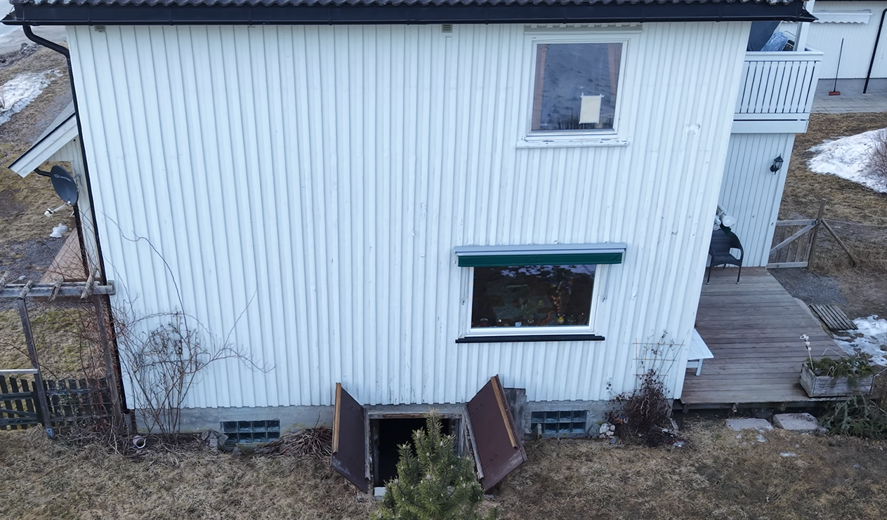 Et utvendig bilde av et hvitt hus med vertikale sider, en kjellerdørinngang og en treterrasse.
