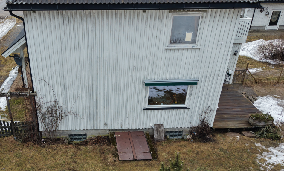 En høyvinkelvisning av et hvitt, vertikalt hjørnet husfasade med to vinduer, en brun kjellerdør og en treterrasse.
