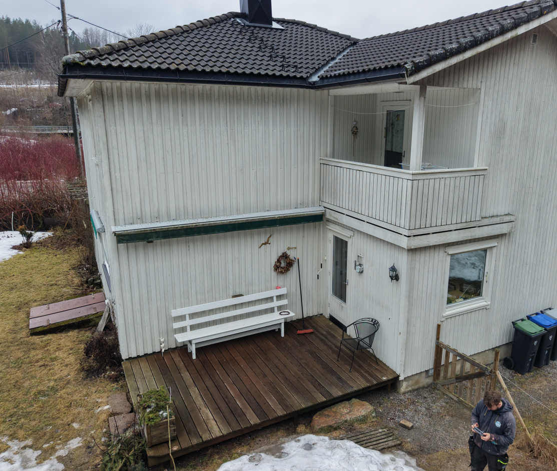 Et toetasjes hvitt trehus med balkong og treterrasse med en hvit benk i en hage med snøflekker.