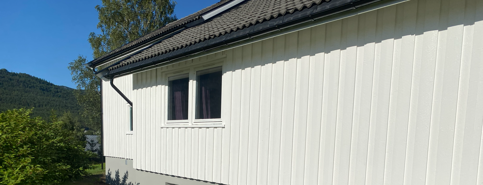 Hvitt trehus eksteriør med vertikal kledning og et mørkt tak, mot en lys blå himmel og fjerne åser.