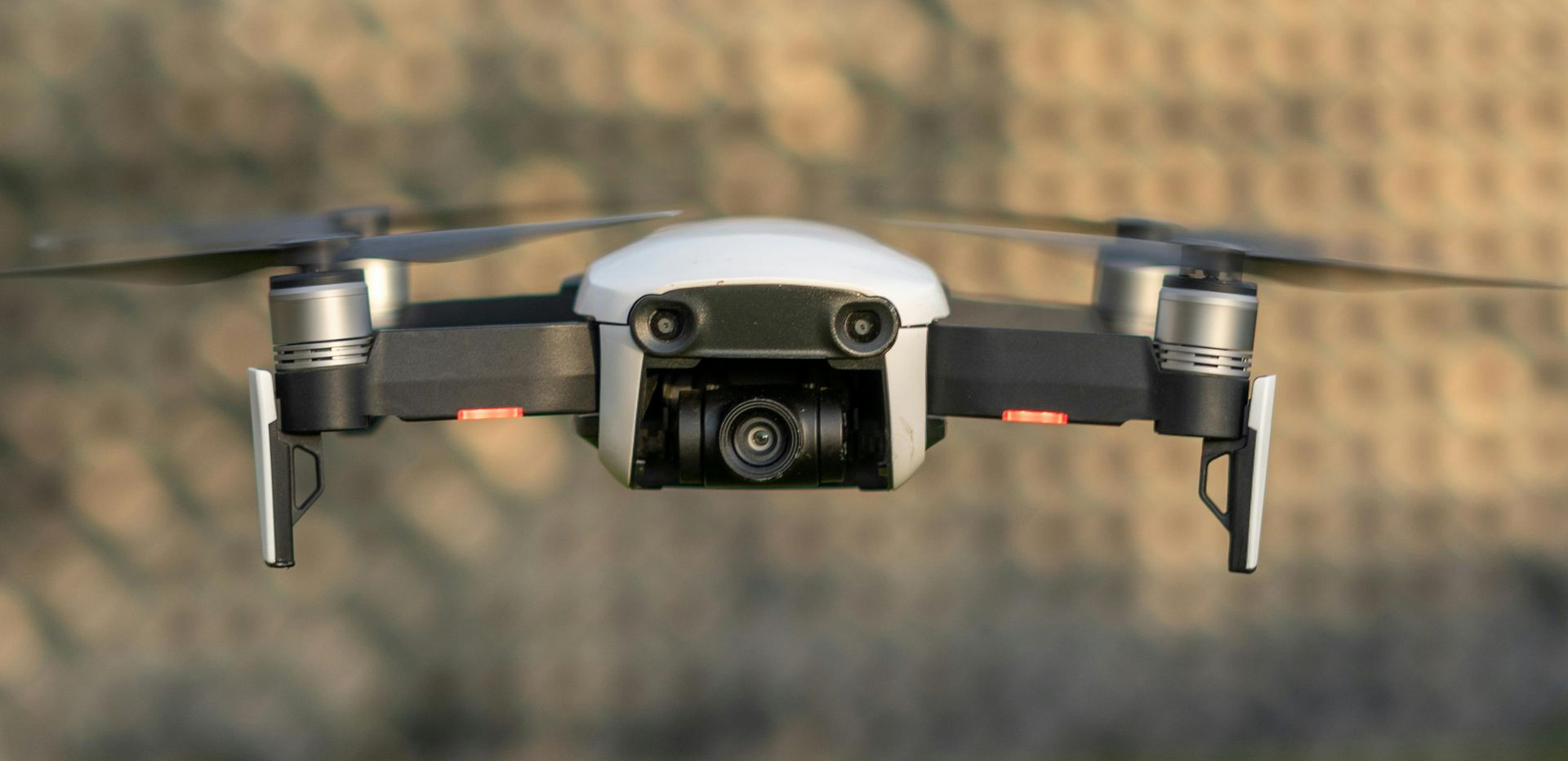 Drone in flight, front view. White and black body with four propellers. Blurred background.