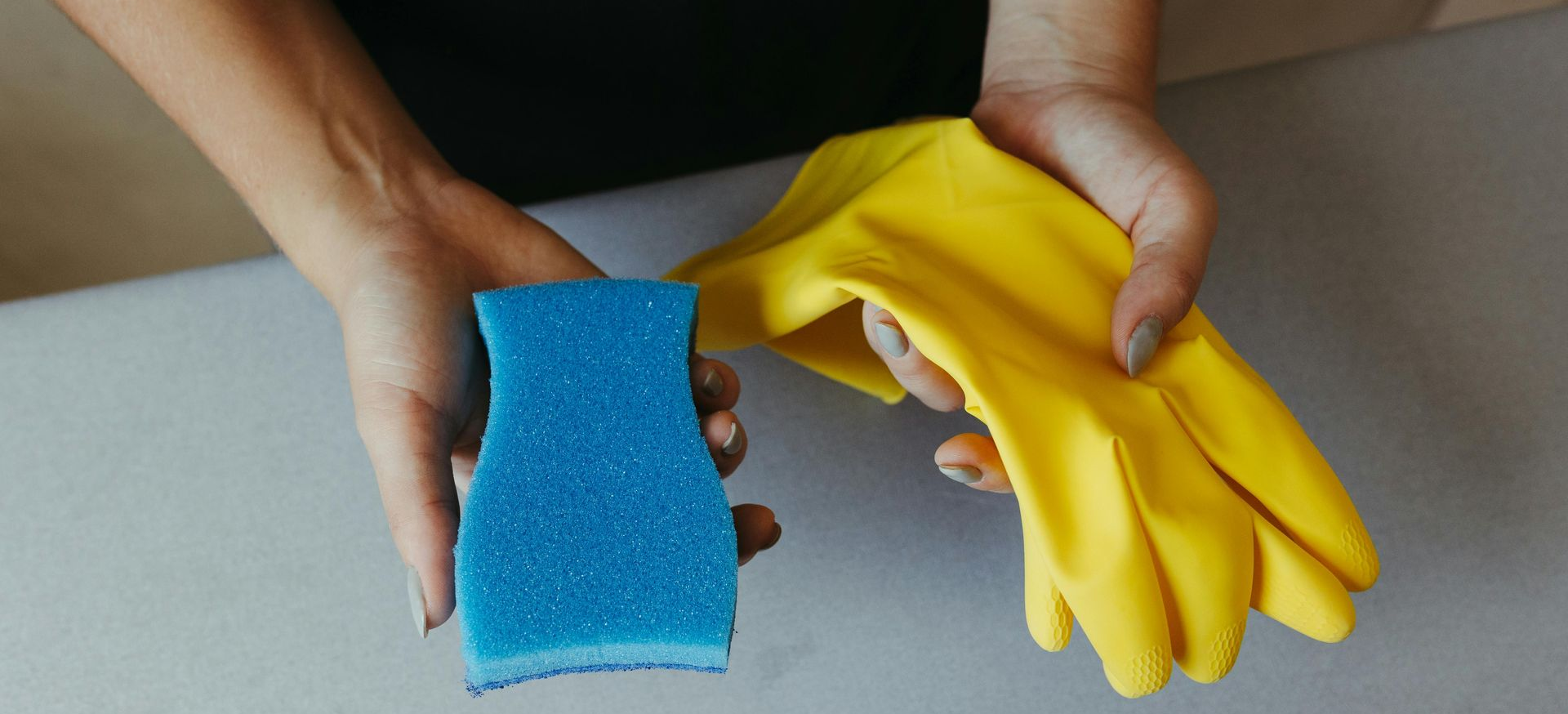 Woman cleaning a kitchen drawer with a blue cloth, wearing a glove; white countertop and cabinets visible.