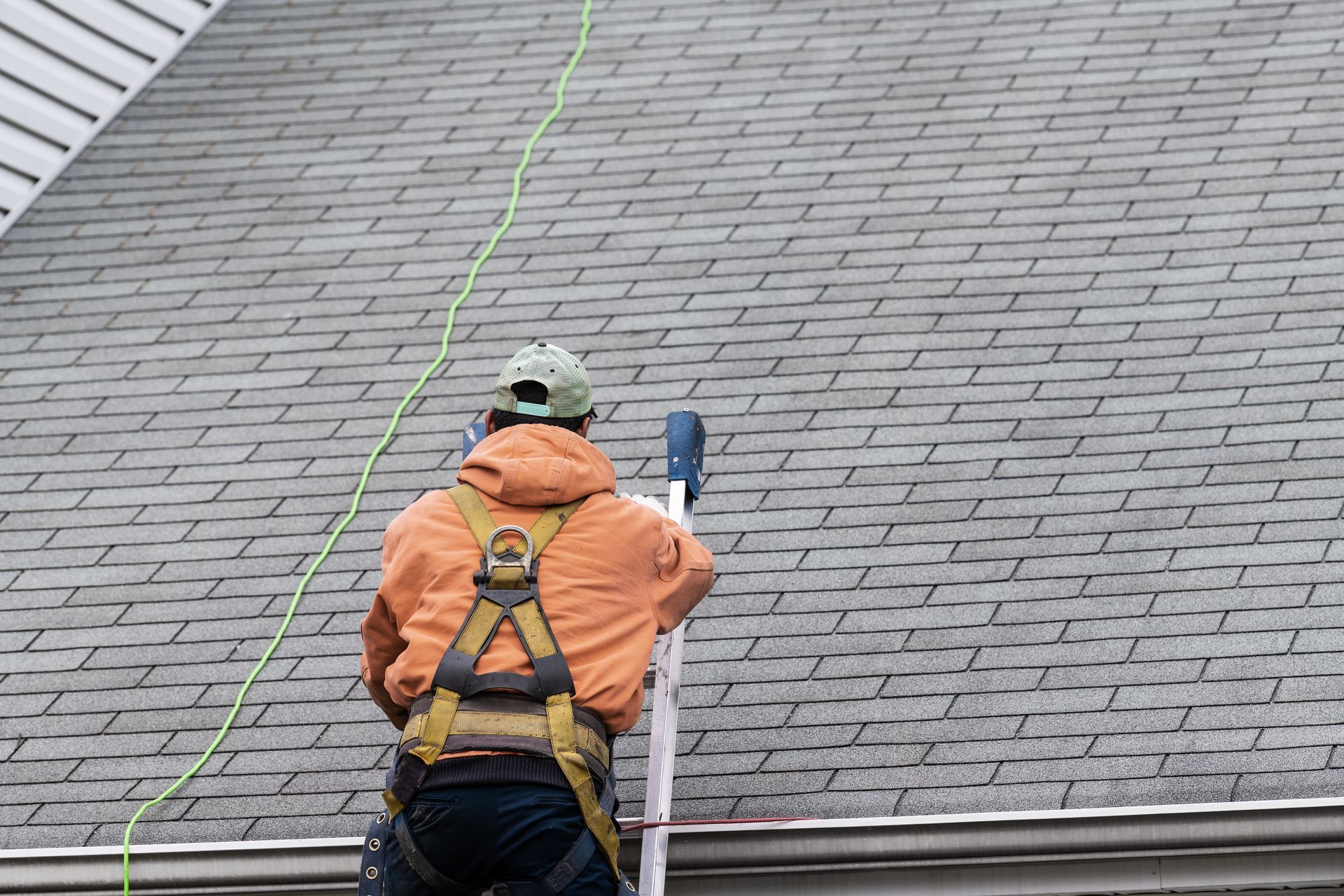 Roofing technician in safety harness climbing ladder onto shingled roof.