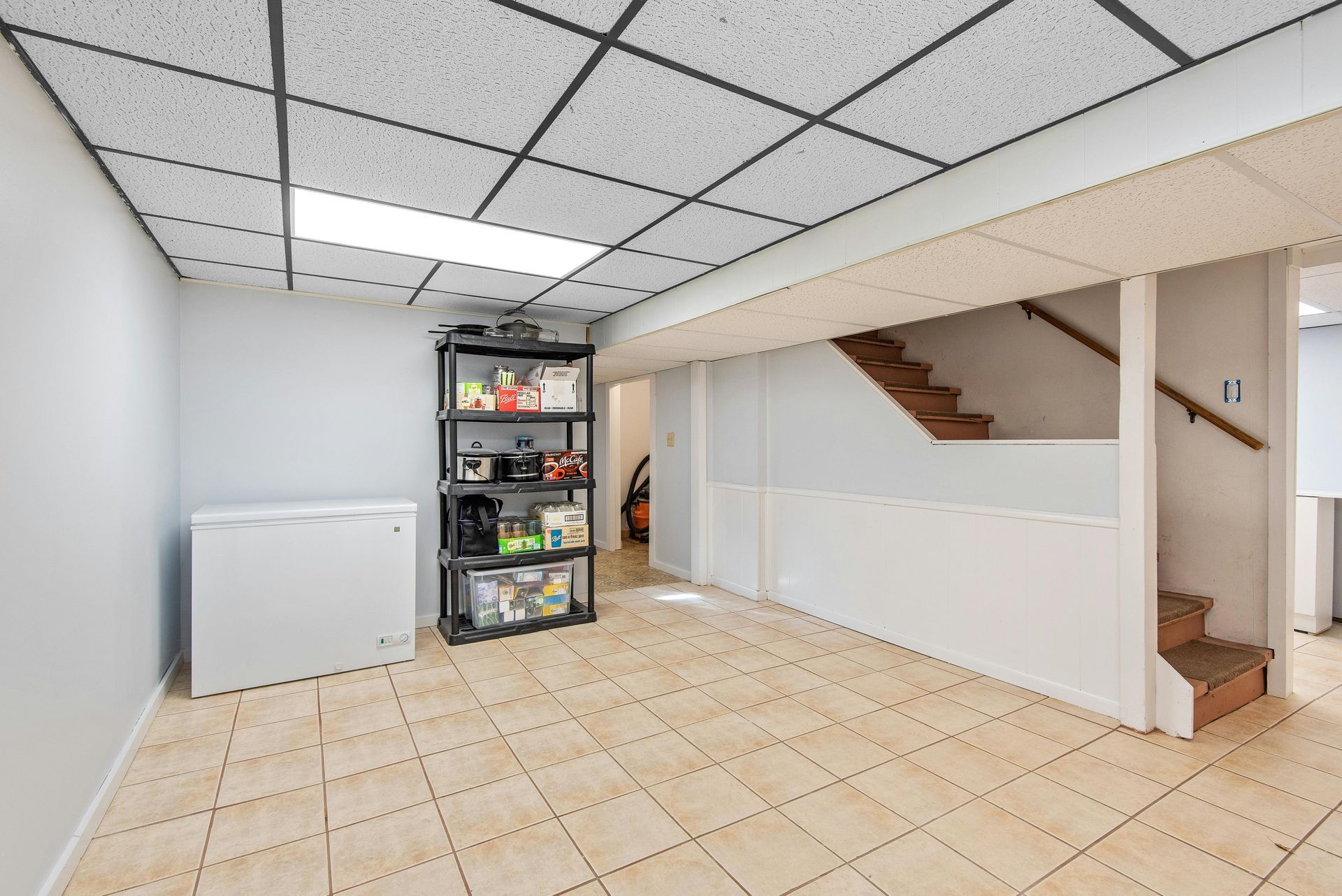 Basement storage area with white walls, tiled floor, and a black shelving unit. Staircase and freezer visible.