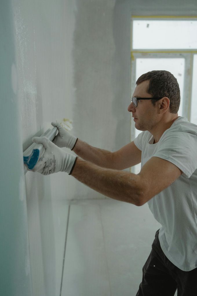 Man in white t-shirt and gloves smoothing compound on a wall with a sanding block.