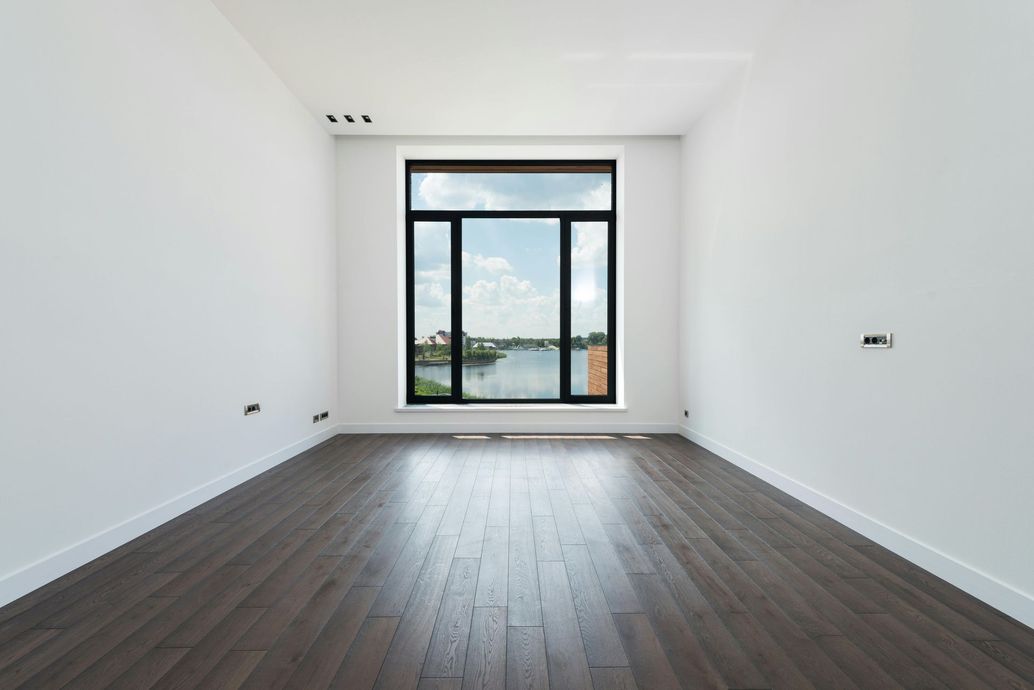 Empty room with dark wood floor, white walls, large window overlooking a body of water.