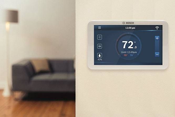 Bosch smart thermostat on beige wall, displaying 72 degrees. Living room scene with a couch and lamp in background.