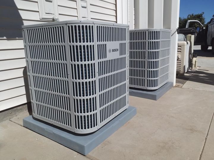 Two gray air conditioning units on concrete pads beside a building.