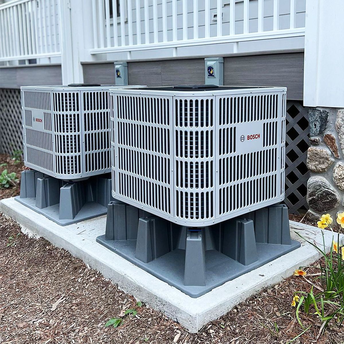 Two gray air conditioning units on concrete pad, mounted beneath a wooden deck.