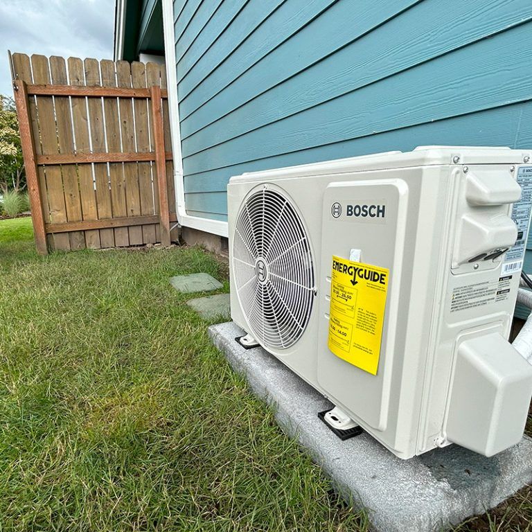 An air conditioner unit on a concrete block next to a blue house and wooden fence on green grass.