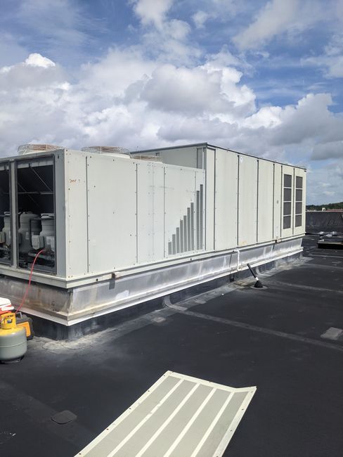 HVAC unit on a flat rooftop under a cloudy sky. The unit is white/gray, and the roof is black.