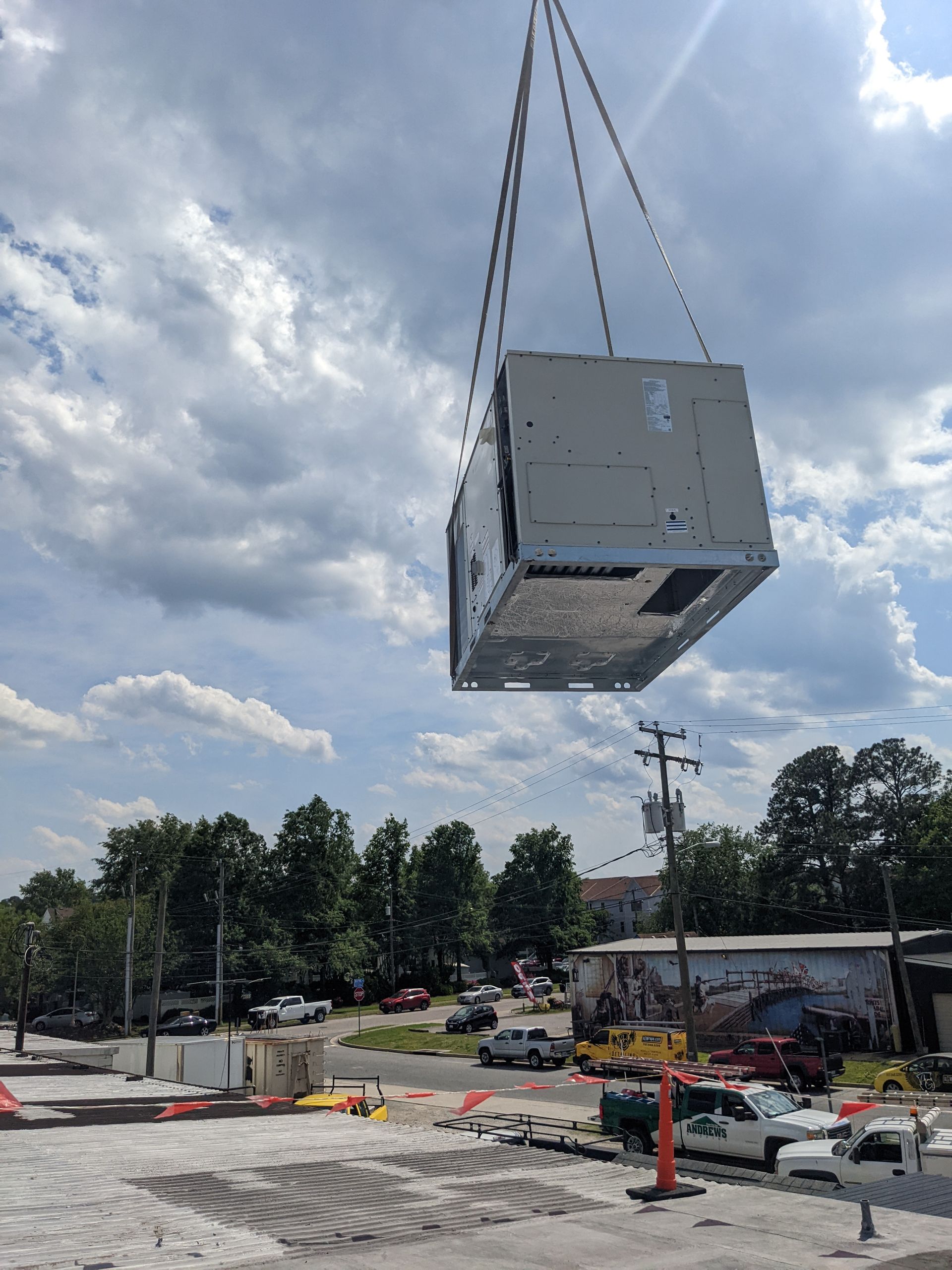 A large HVAC unit is lifted by a crane against a cloudy sky. Construction site below.