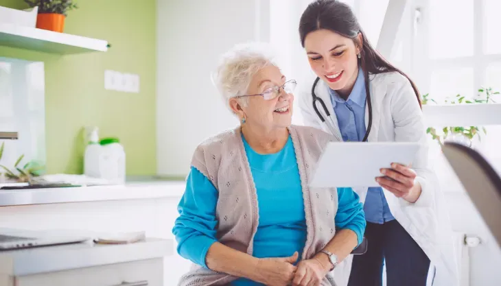 A doctor is talking to an elderly woman while looking at a tablet.