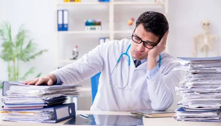 A doctor is sitting at a desk with a stack of papers.