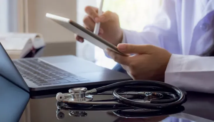 A doctor is using a tablet computer while sitting at a desk with a laptop and a stethoscope.