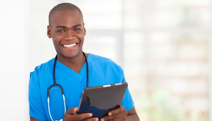 A man in a blue scrub is holding a tablet and smiling.