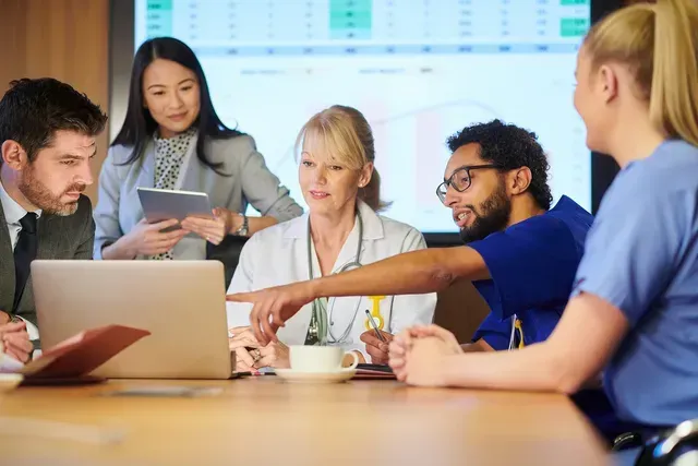 A group of doctors and nurses are sitting around a table looking at a laptop.