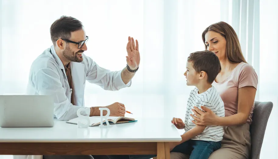 A doctor is giving a child a high five while sitting at a table with a woman and child.