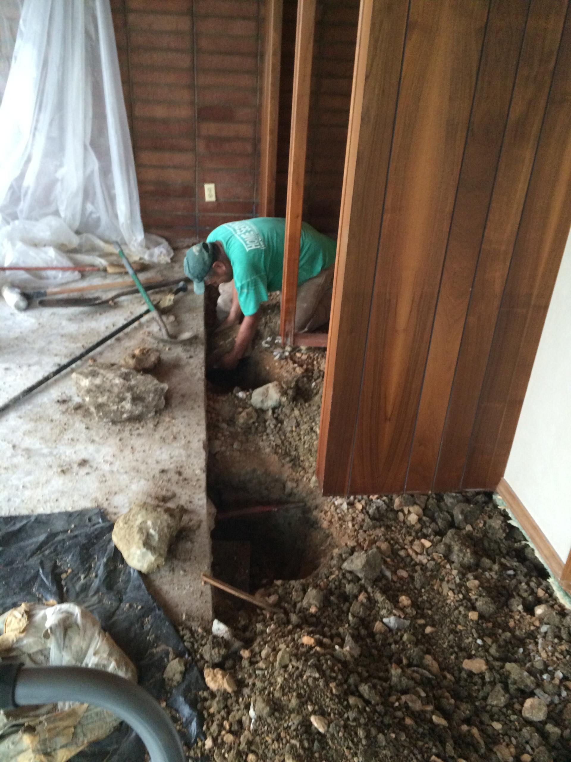 Person digging in dirt floor between wooden walls, in a room. Debris and equipment surround them.