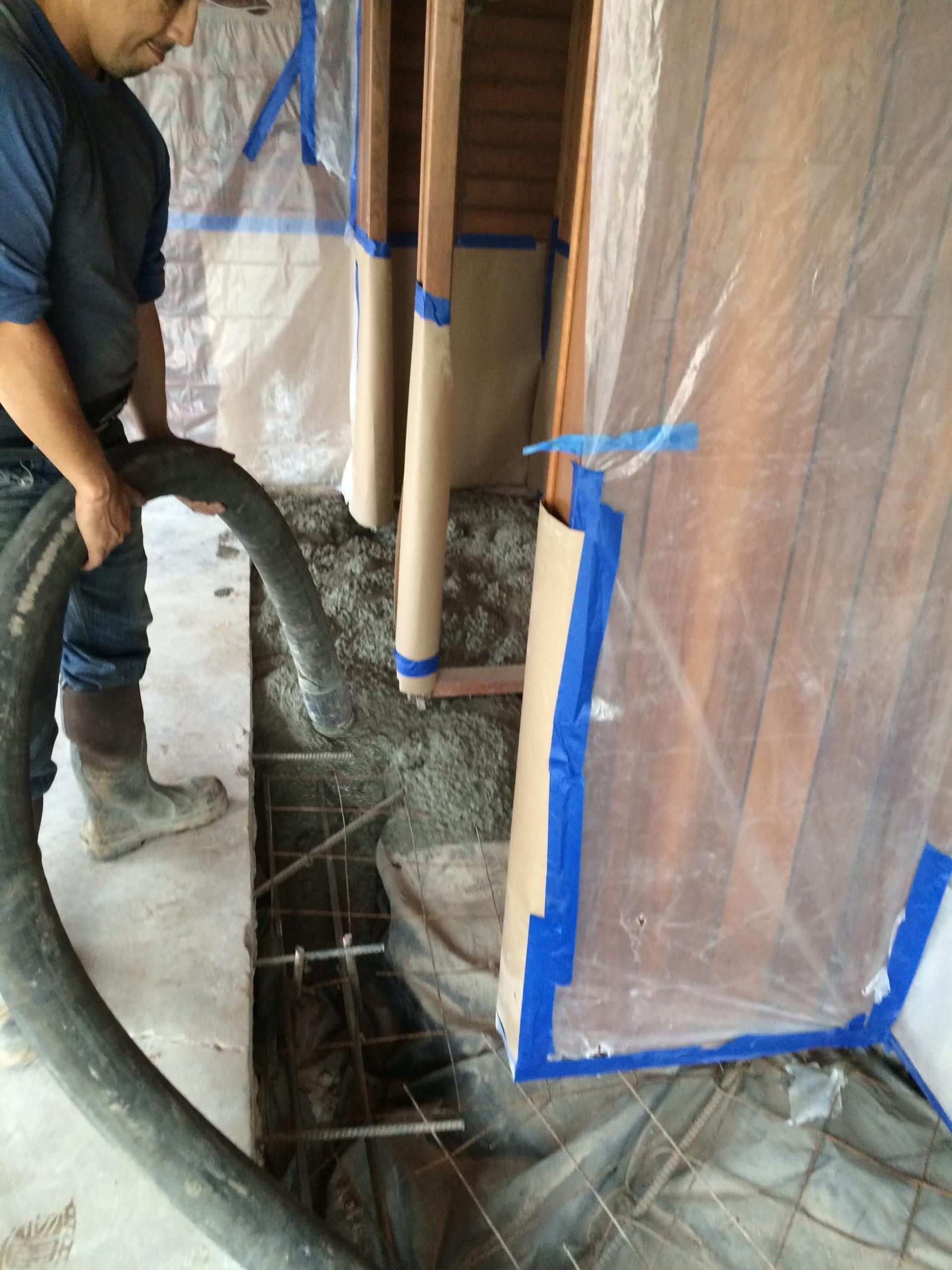 Man pouring concrete from a hose into a trench with rebar, plastic sheeting protects wooden walls.