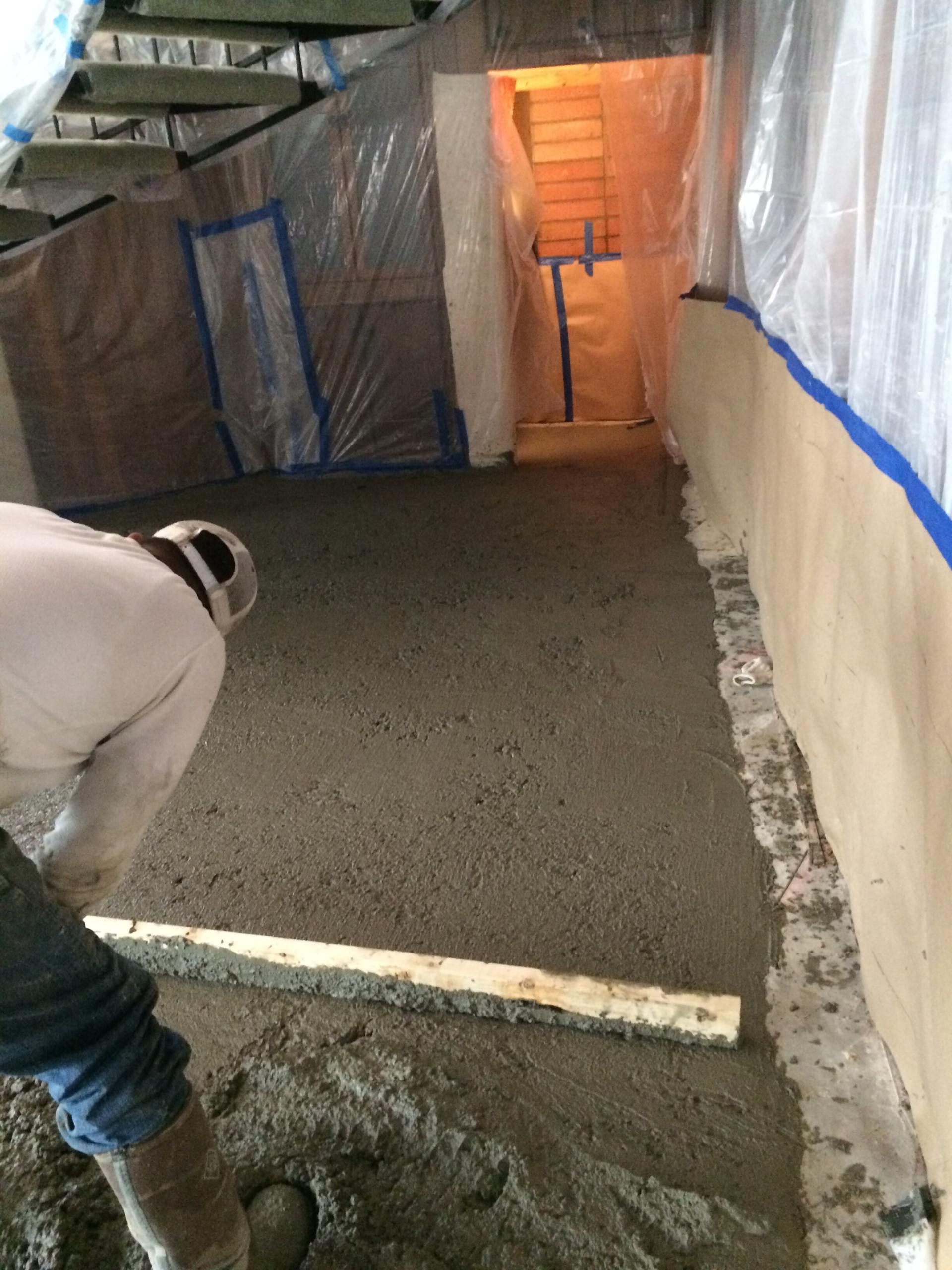 Construction worker smoothing concrete floor with a wooden plank in an enclosed area; plastic sheeting covers walls.