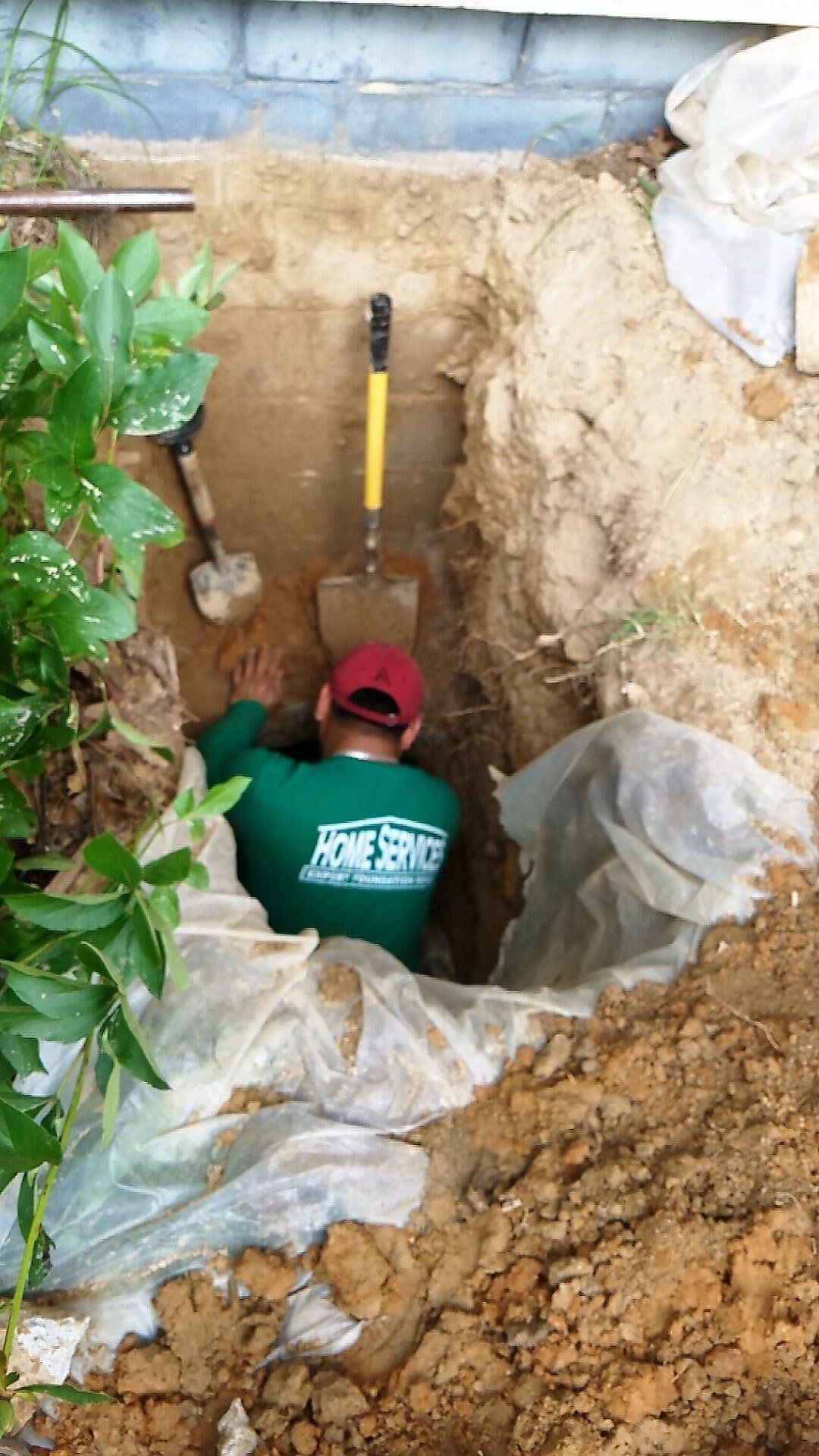 Person in a trench, digging near a house. He wears a green shirt and red hat. Tools lay beside him.