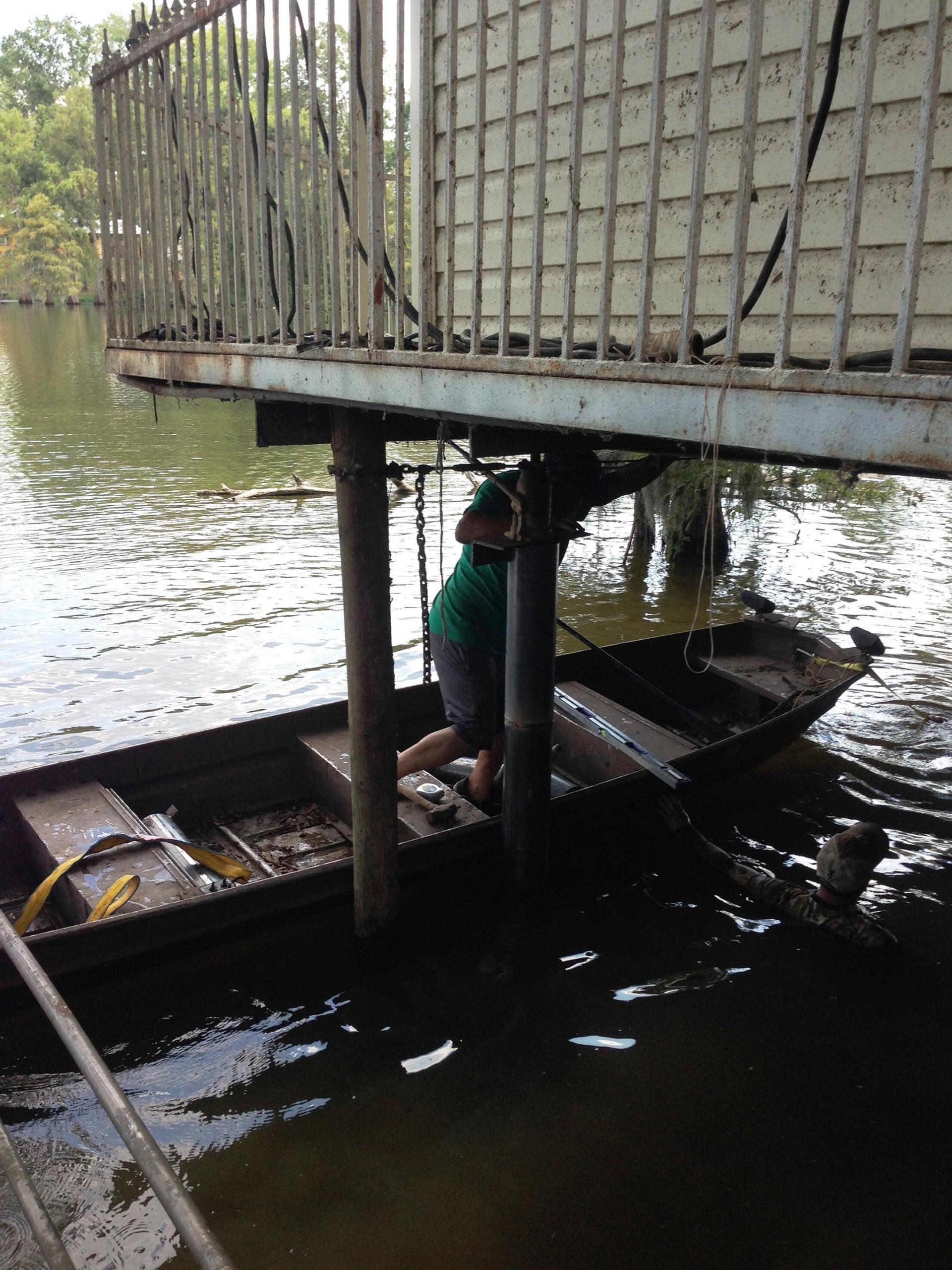 A person in a green shirt stands in a small boat under a dock, near water and a railing.