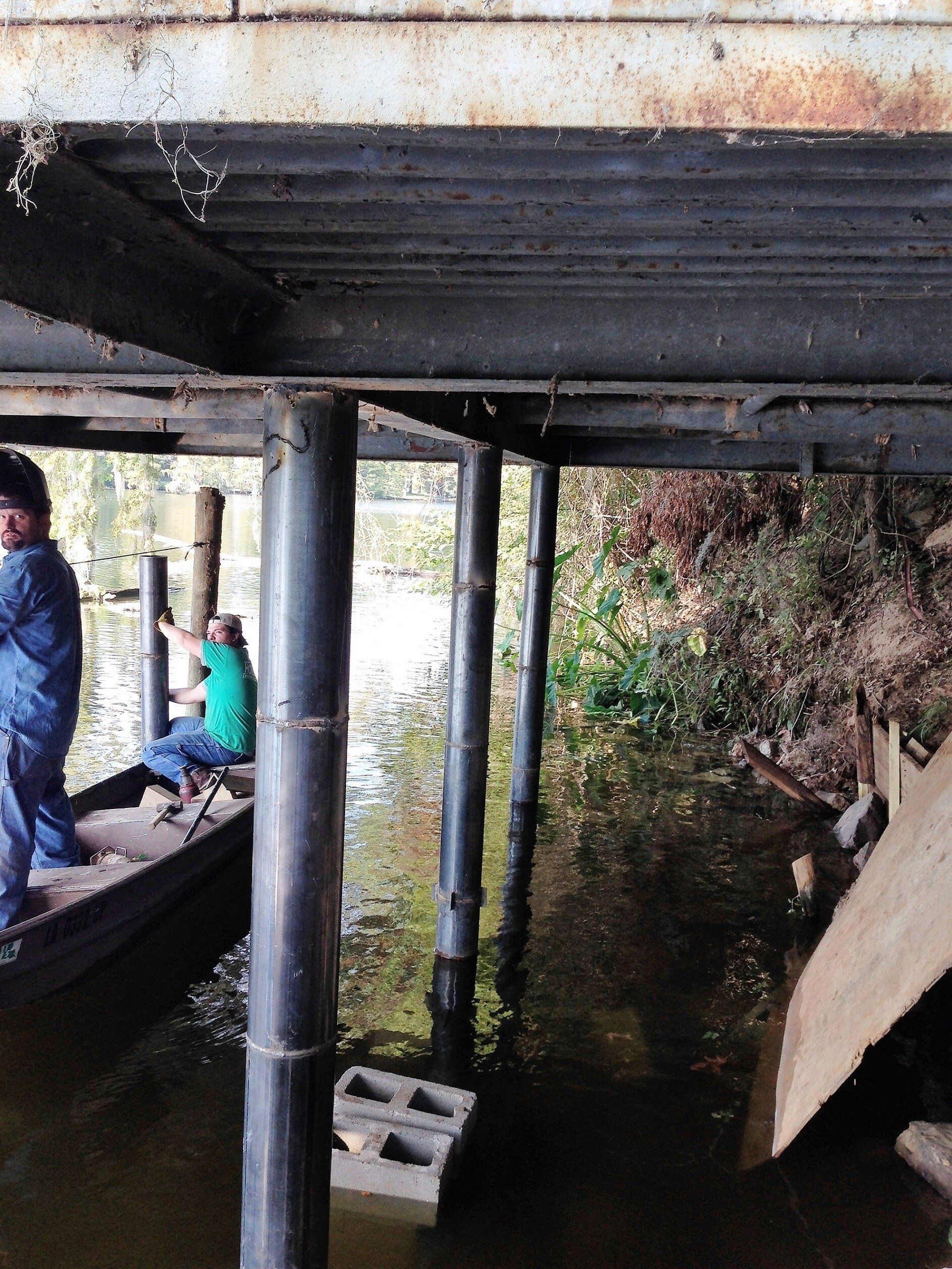 Men on a boat near bridge supports in water. Rusty metal beams overhead. Vegetation, water.