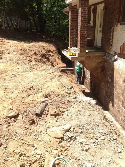 Construction site: worker by a house foundation, digging in dirt. Brown earth and a red brick house.