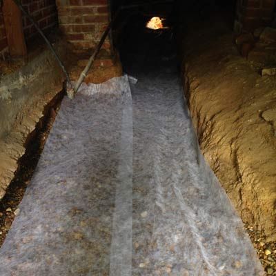 Gravel-filled trench covered with white fabric, in a dark, brick-walled crawl space.