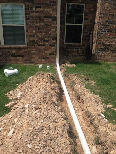 A trench with a white pipe runs along a brick wall, with windows on the left and right.
