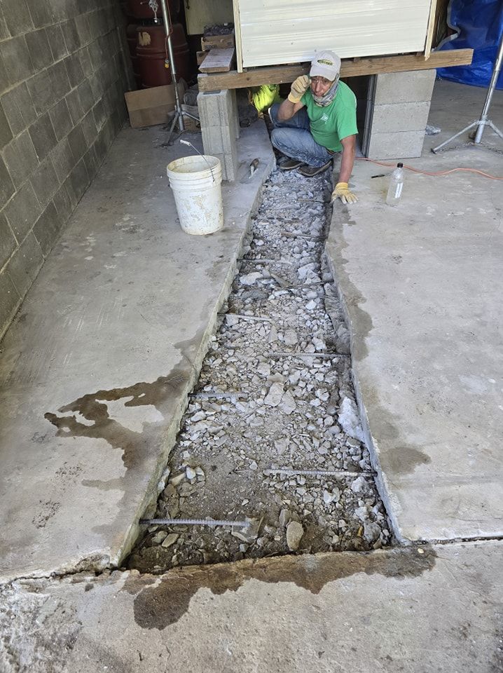 Man in a green shirt repairing a concrete floor, digging a trench, with rebar in place. Outdoors in a garage.