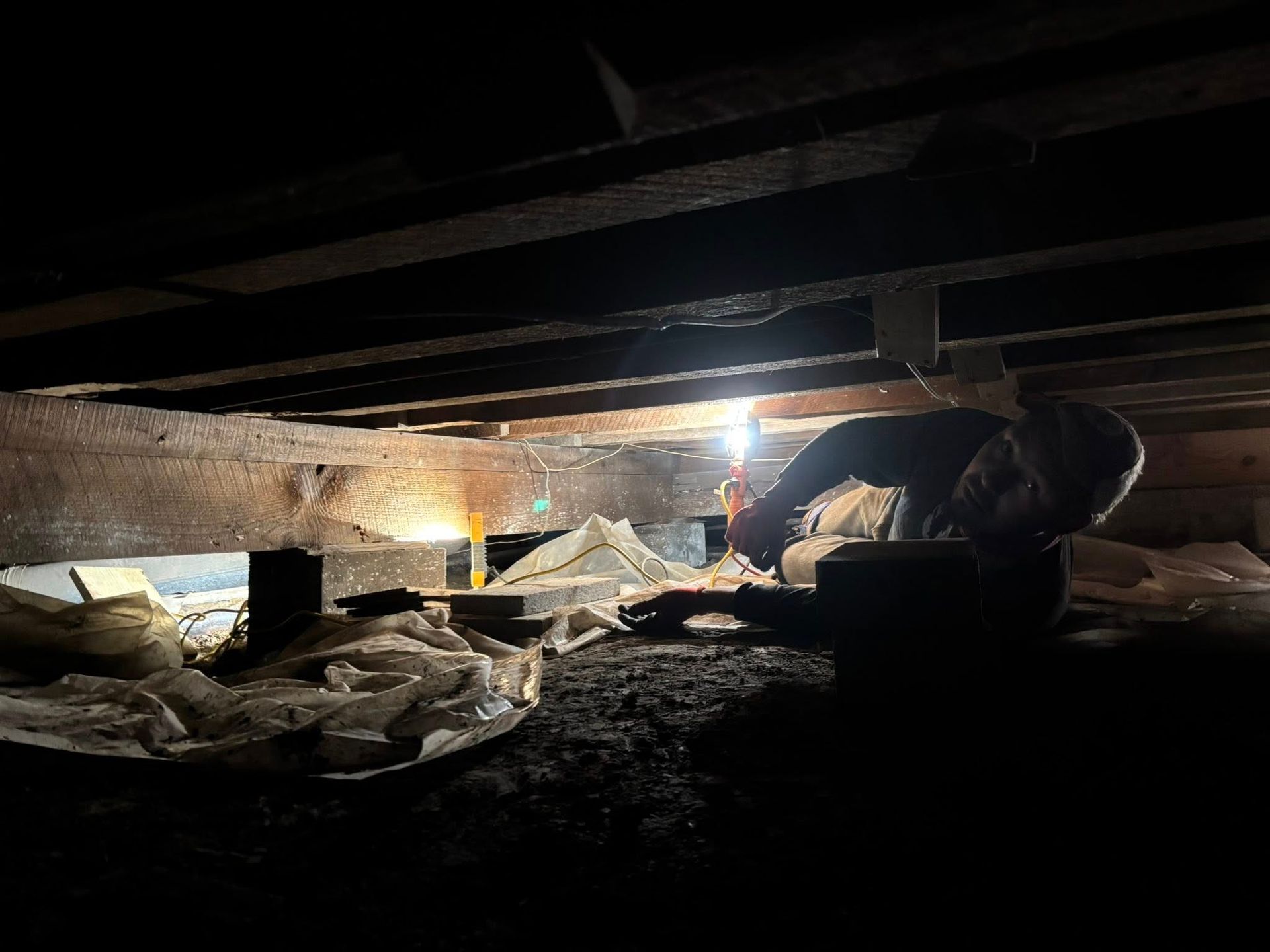 Person examines the underside of a dark crawl space, illuminated by a light.