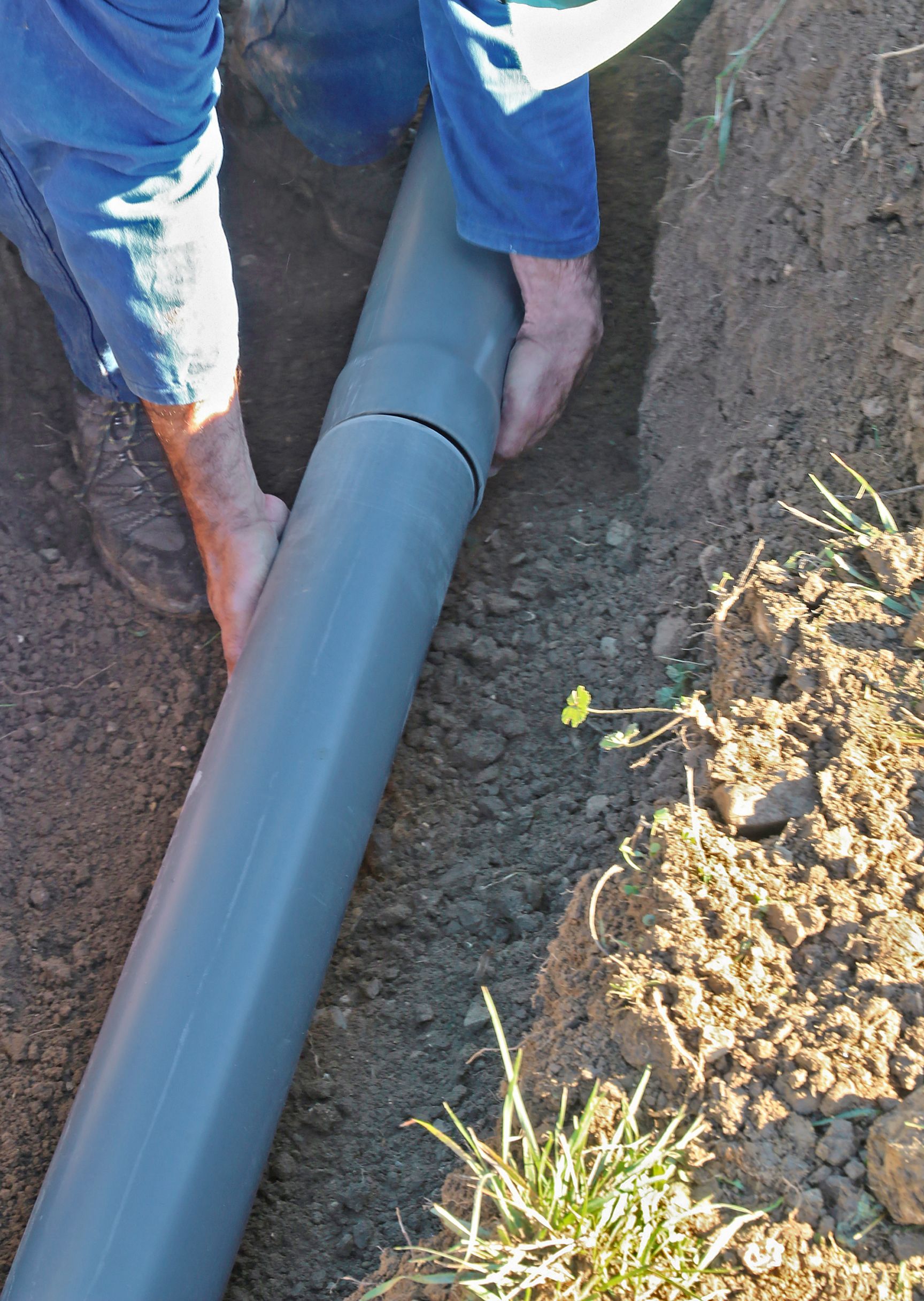 A trench dug in a yard with retaining walls, a backhoe in the background, and leaves scattered throughout.