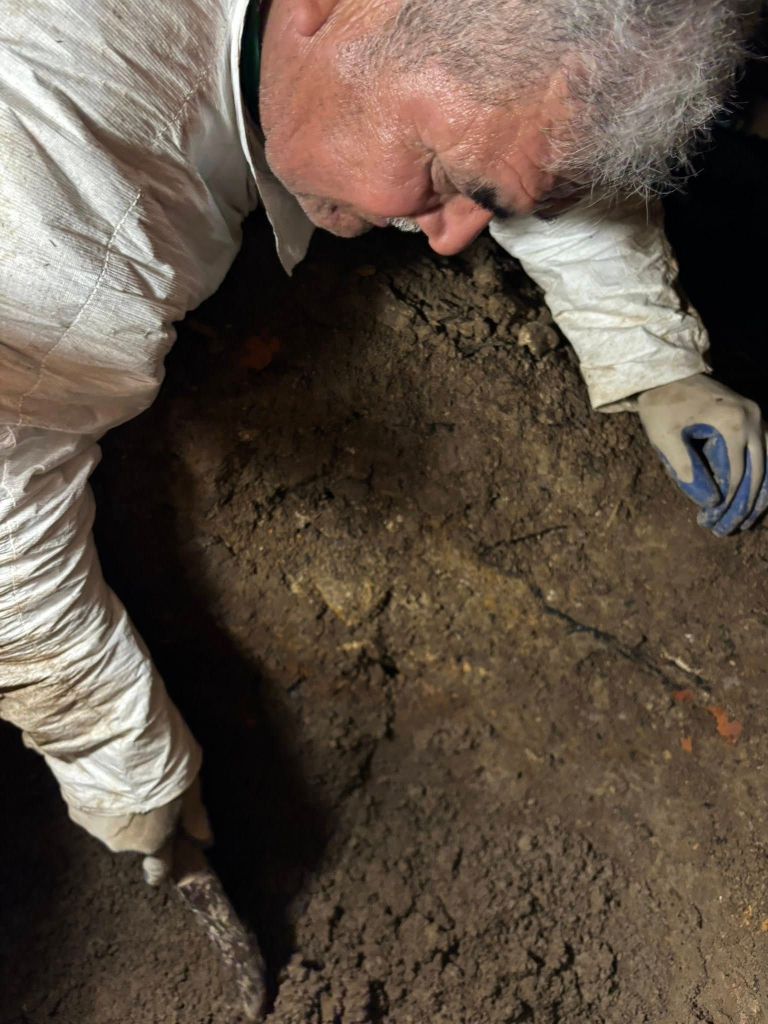 Person in white protective suit, examining soil with a trowel.