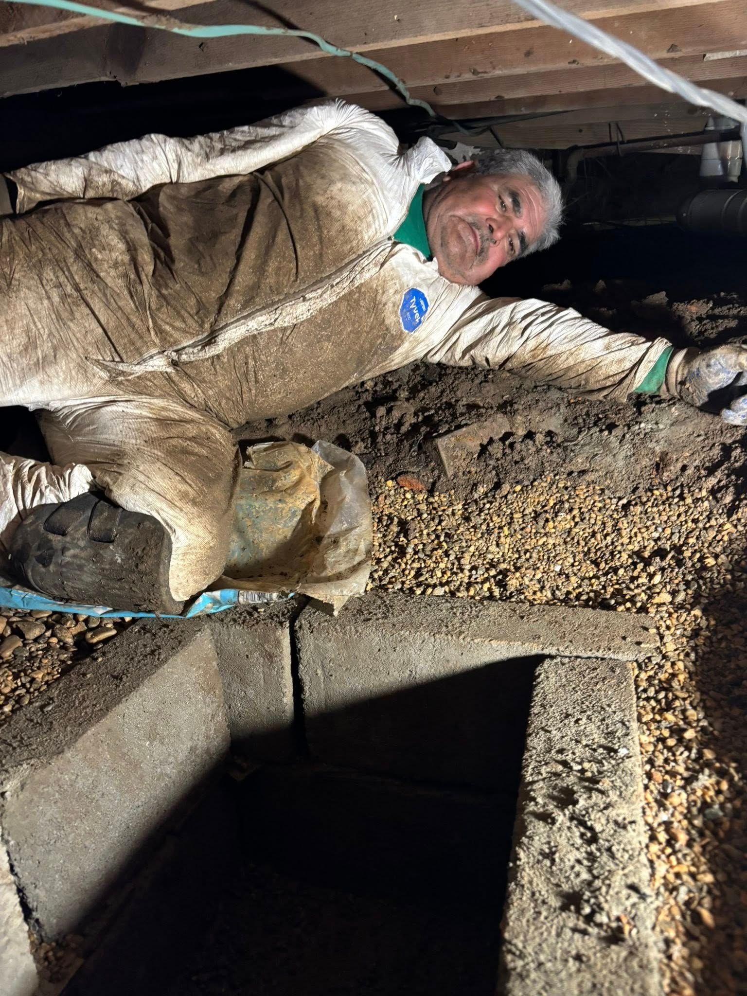 Man in protective suit working under a house with dirt and debris.