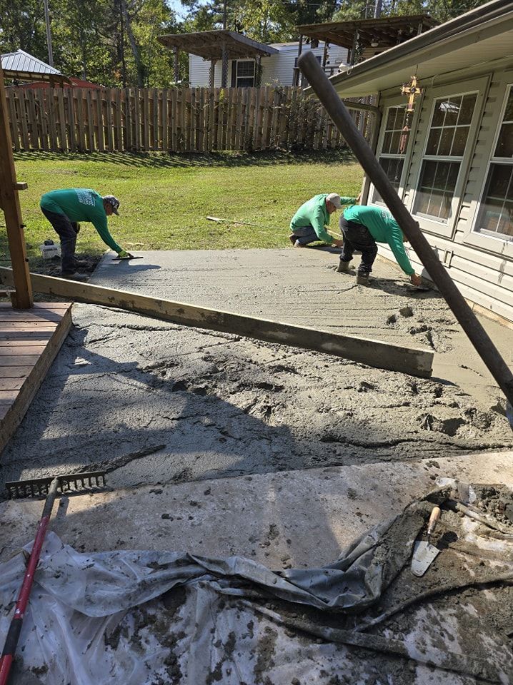 Three workers in green shirts smoothing wet concrete on a patio next to a house.