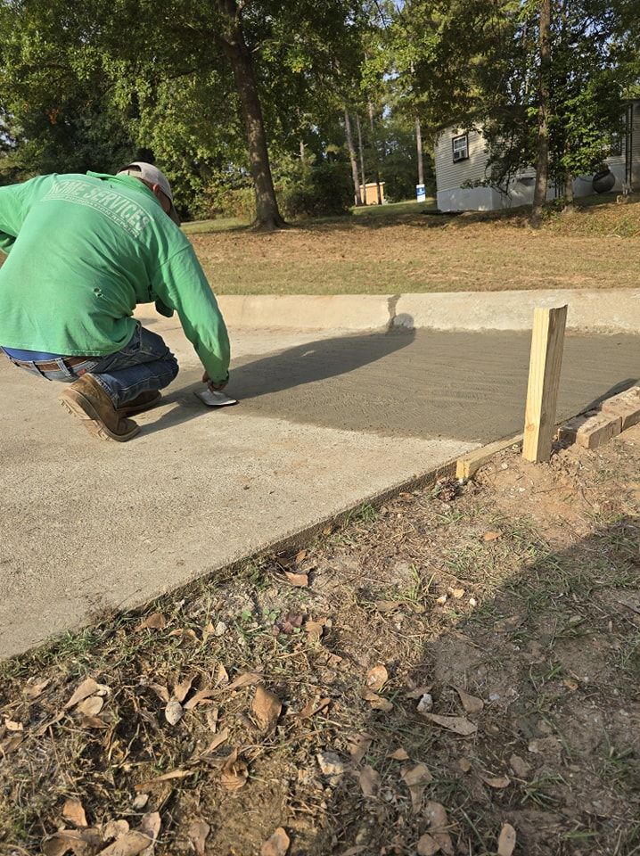 Person in green shirt smoothing wet concrete on a sidewalk, wooden stakes mark the border.