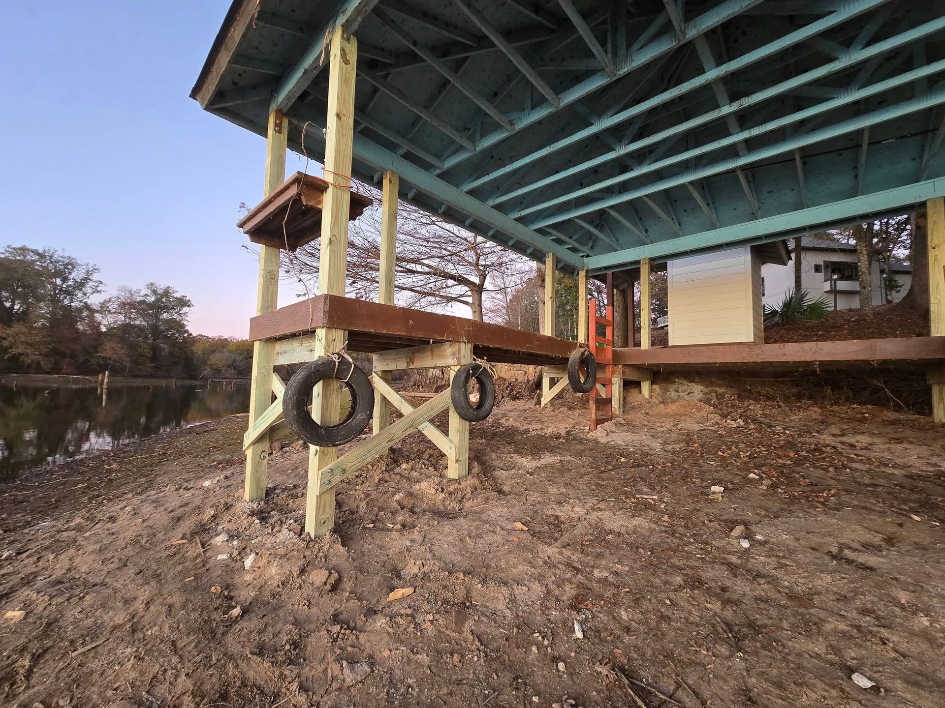 A wooden lakeside dock with tires hanging beneath a roofed structure, dirt foreground, and trees in the distance.