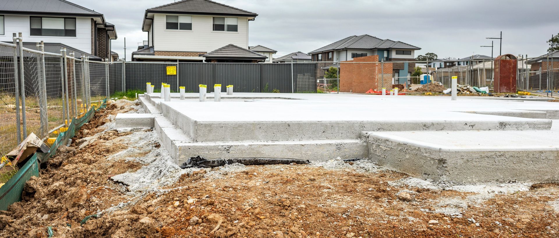 Person in green shirt smoothing wet concrete on a sidewalk, wooden stakes mark the border.