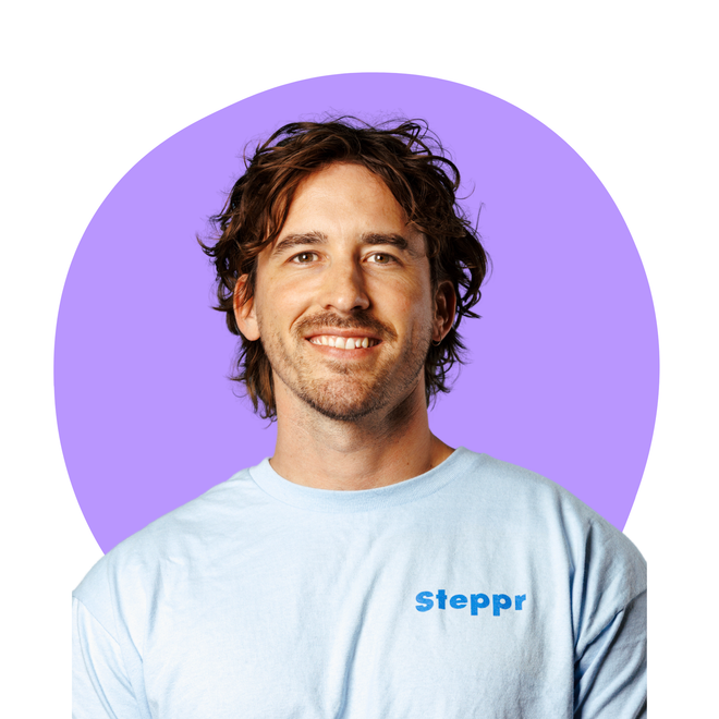 A person with wavy brown hair and light stubble smiles while wearing a light blue