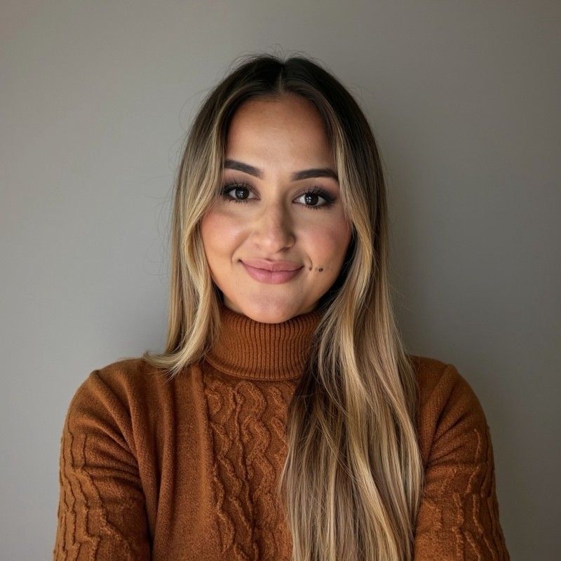 A person with long, two-toned blonde and brown hair wearing a brown cable-knit turtleneck sweater against a gray background.